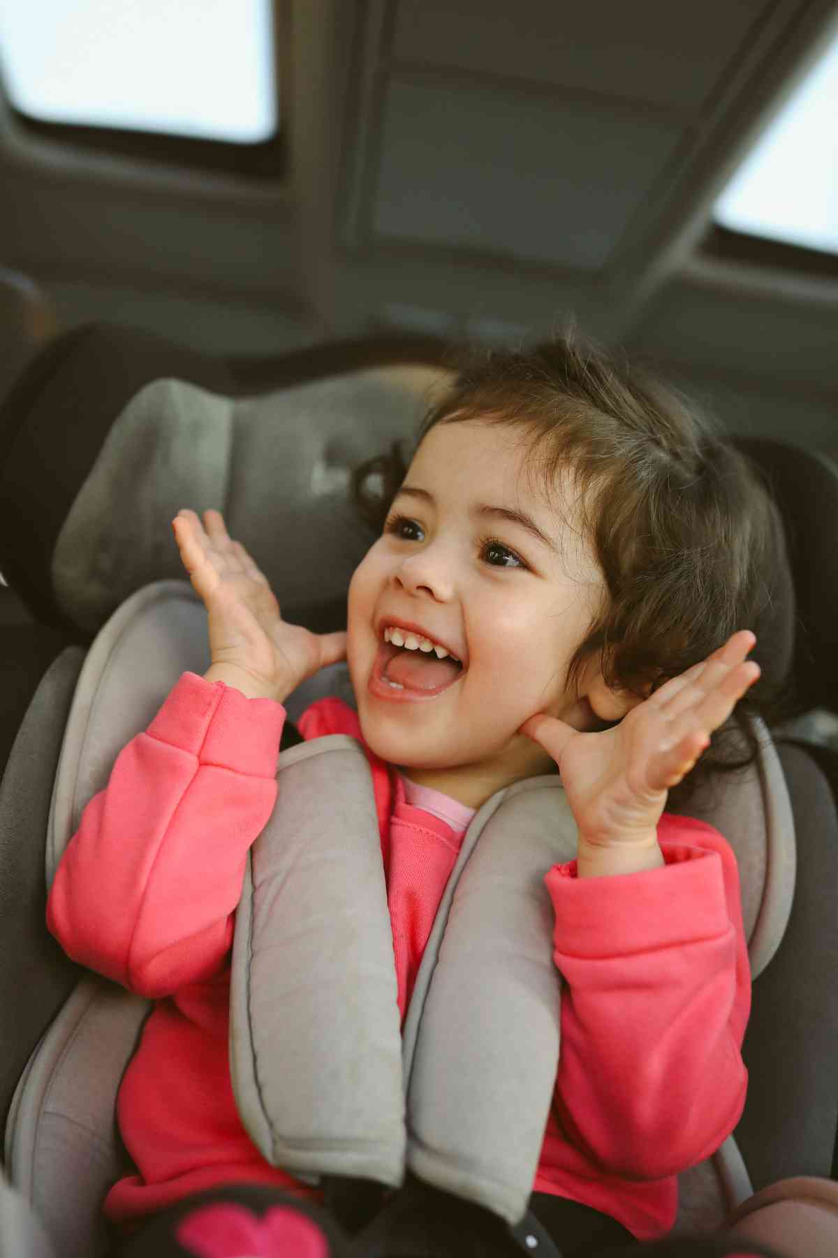 A young child with short dark hair, wearing a pink jacket, smiling and sitting in a car seat with hands raised near their face.