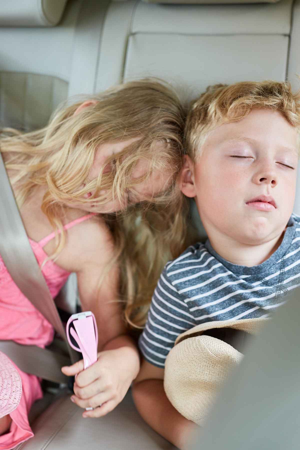 Two children are sleeping in a car's backseat, both wearing seatbelts.