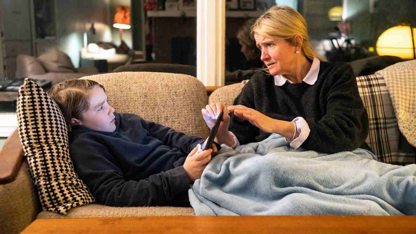 An adult and a child sit on a couch; the adult gestures while talking to the child, who is holding a tablet.