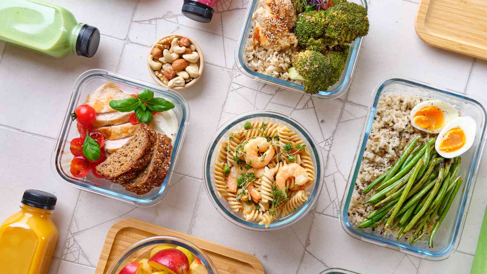 An assortment of healthy meals in glass containers, including salad, pasta with shrimp, boiled eggs, green beans, and rice, arranged on a tiled surface with bottles of juice and nuts.