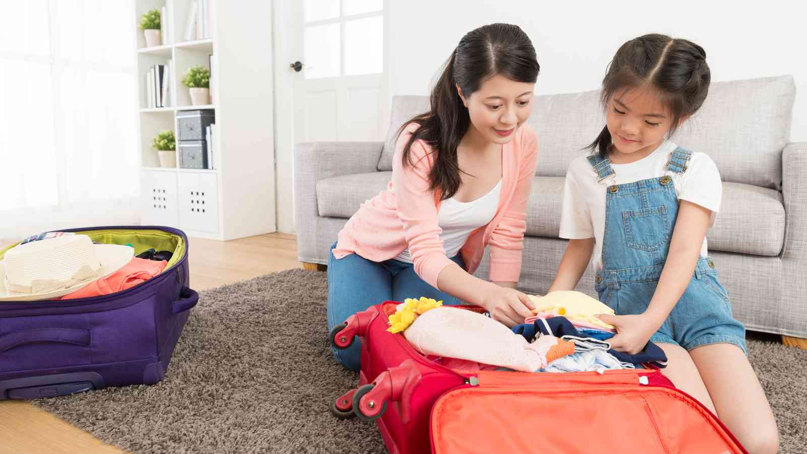 A mom is helping her daughter pack clothes into a red suitcase.