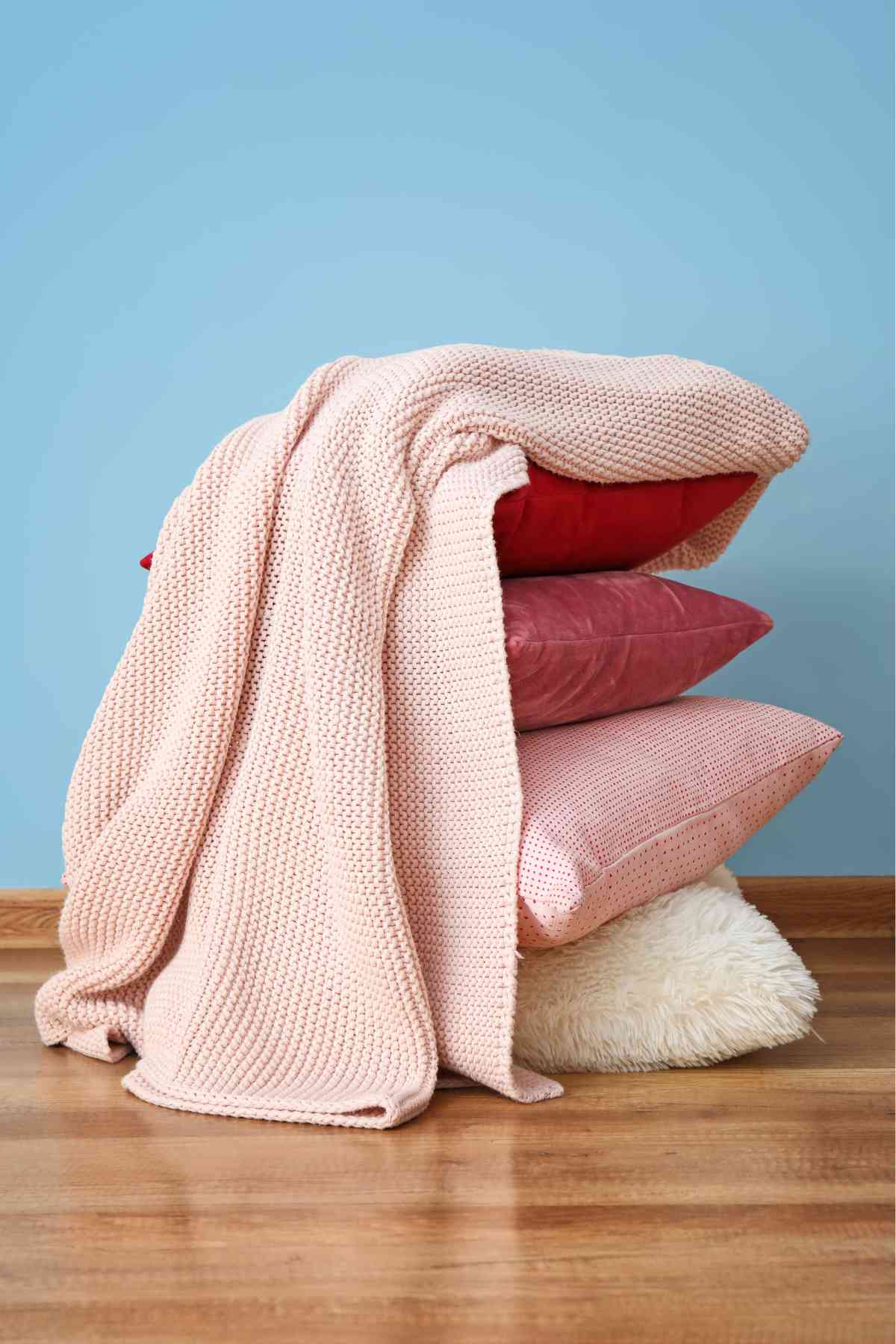 A stack of pillows with red and pink cases, topped with a folded pale pink knitted blanket.