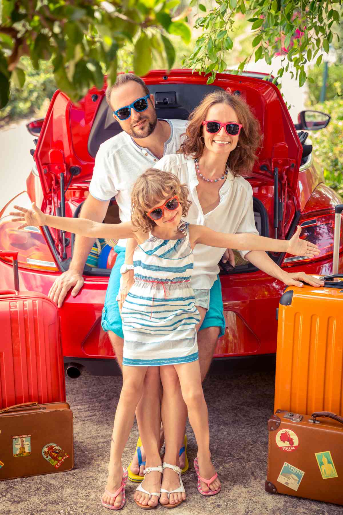 A smiling family of three wearing sunglasses pose in front of an open car trunk with luggage around them.