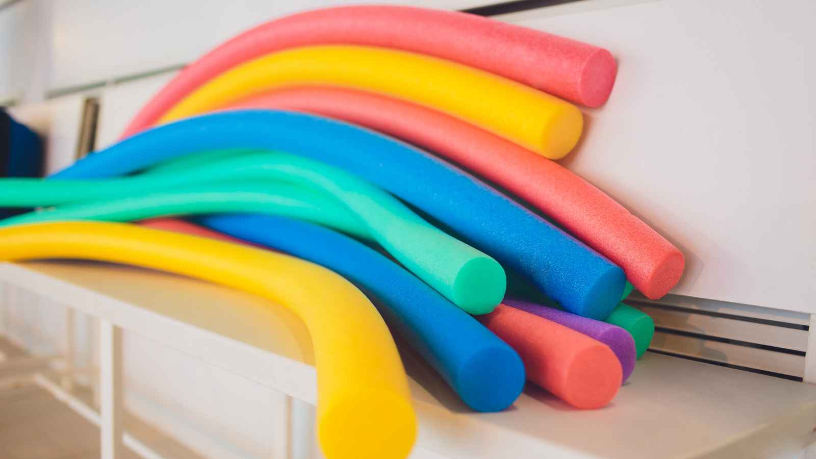 A stack of colorful pool noodles, including yellow, pink, blue, green, and purple, is arranged on a white surface.