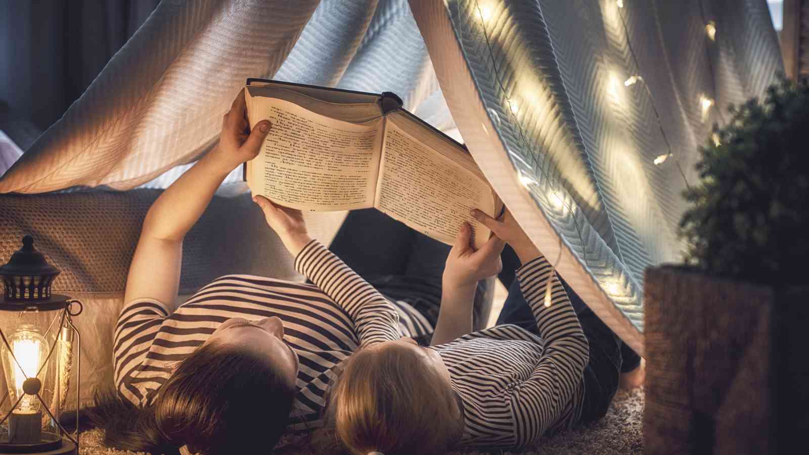 A mom and her child reading a book together under a blanket fort illuminated by string lights and a lantern.