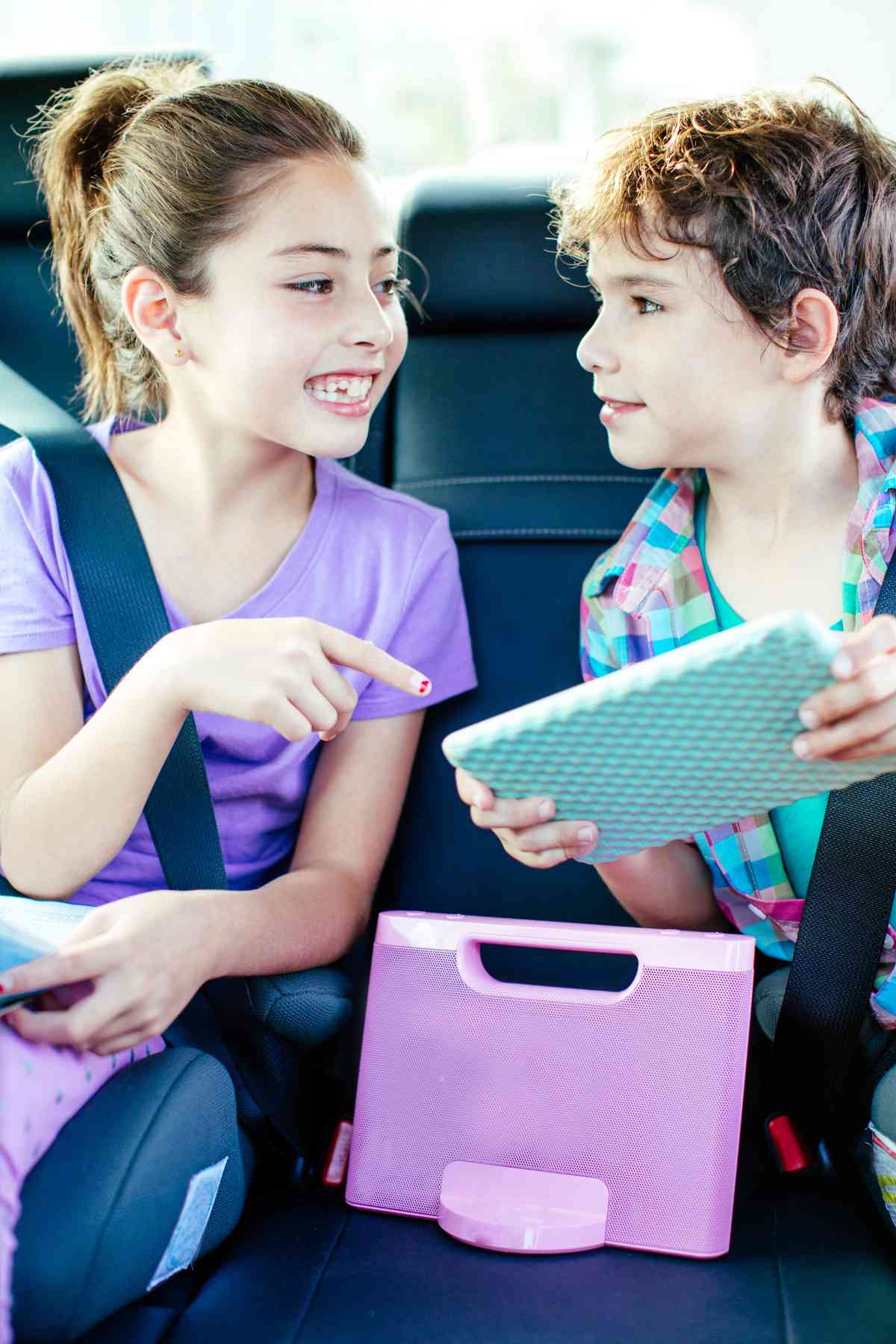 Two children, a girl and a boy, are sitting in the backseat of a car, chatting and interacting with a tablet.