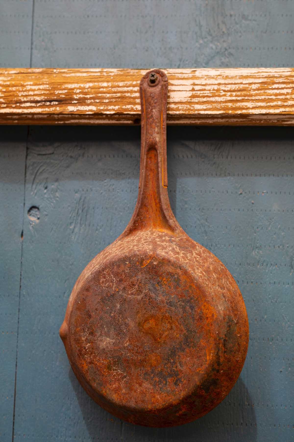 Rusty cast iron skillet hanging on a wooden beam against a blue wall.