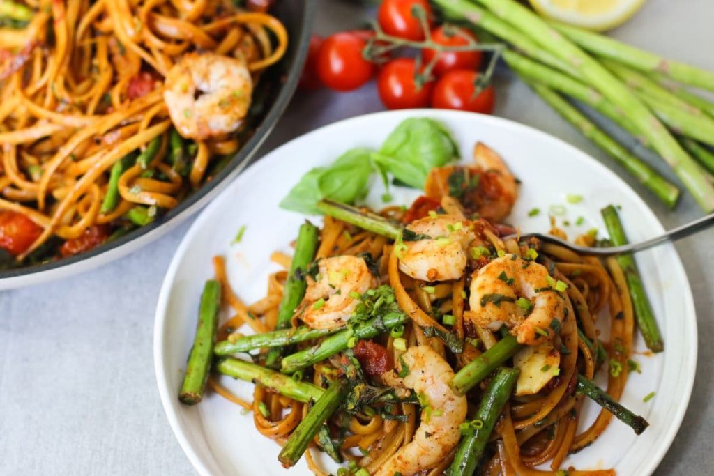 A plate of shrimp tomato basil pasta with asparagus, and herbs, next to a fork.