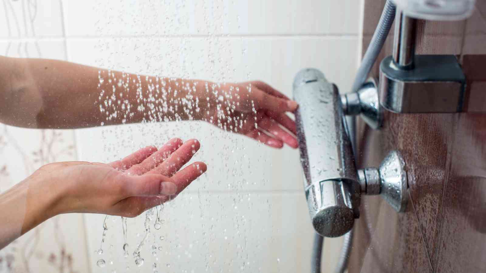 Hands are adjusting the water temperature from a shower head, with water running over one hand under the shower.