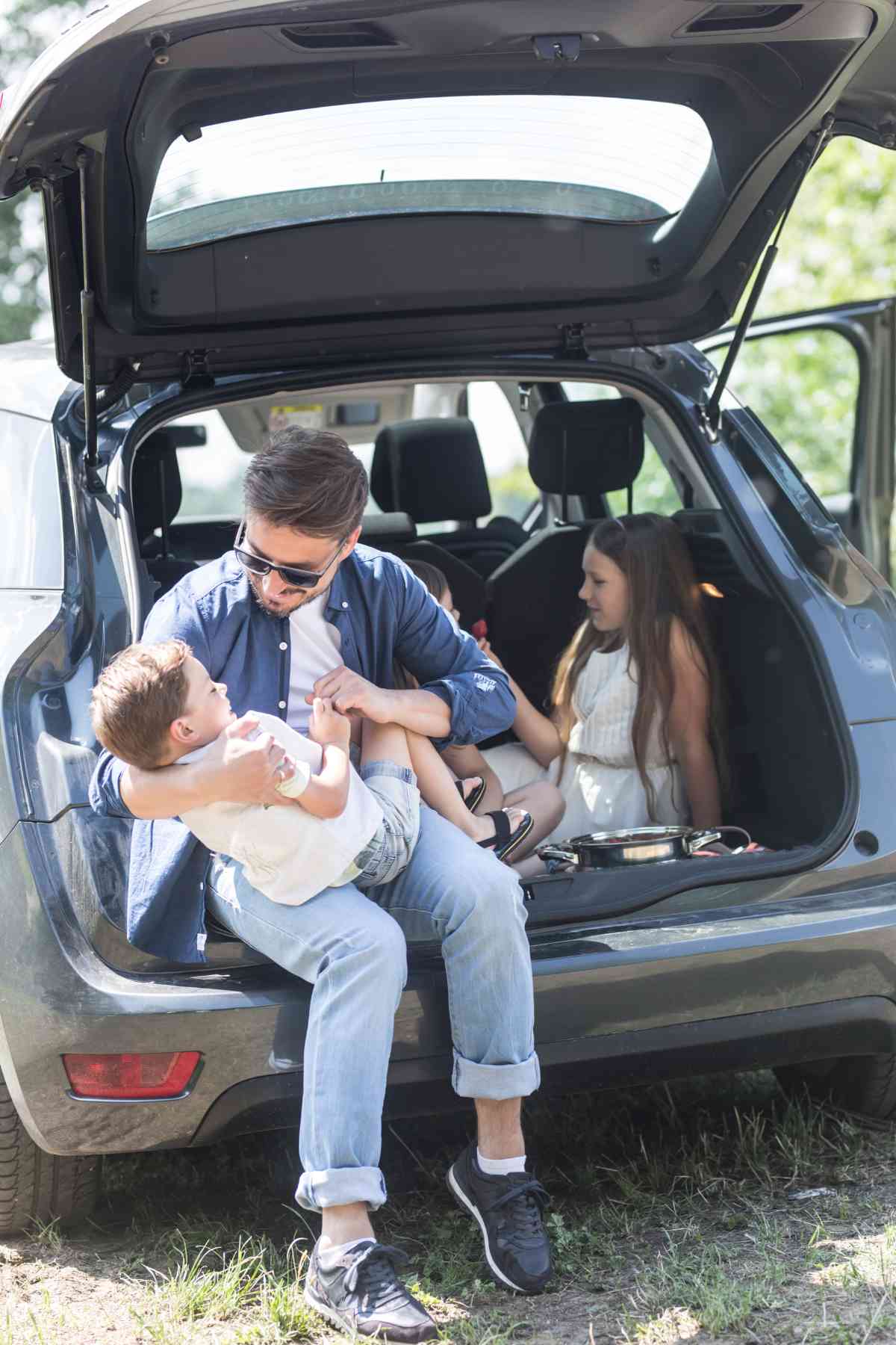 A man sits on the edge of an open car trunk, holding a young boy, while a girl sits inside the trunk.