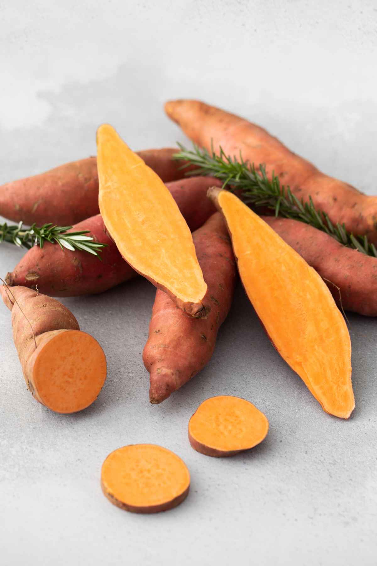 A cluster of whole and sliced sweet potatoes lies on a gray surface, with a sprig of rosemary placed among them.
