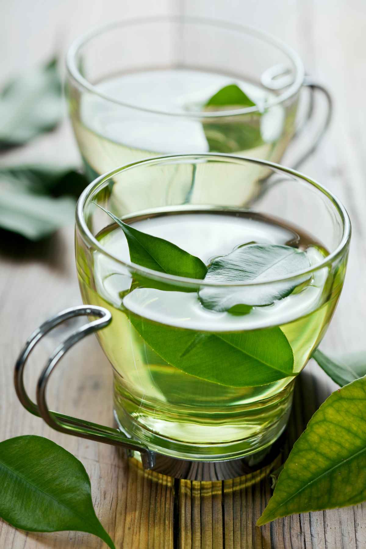 Two clear glass cups filled with green tea, each garnished with green leaves, placed on a wooden surface.