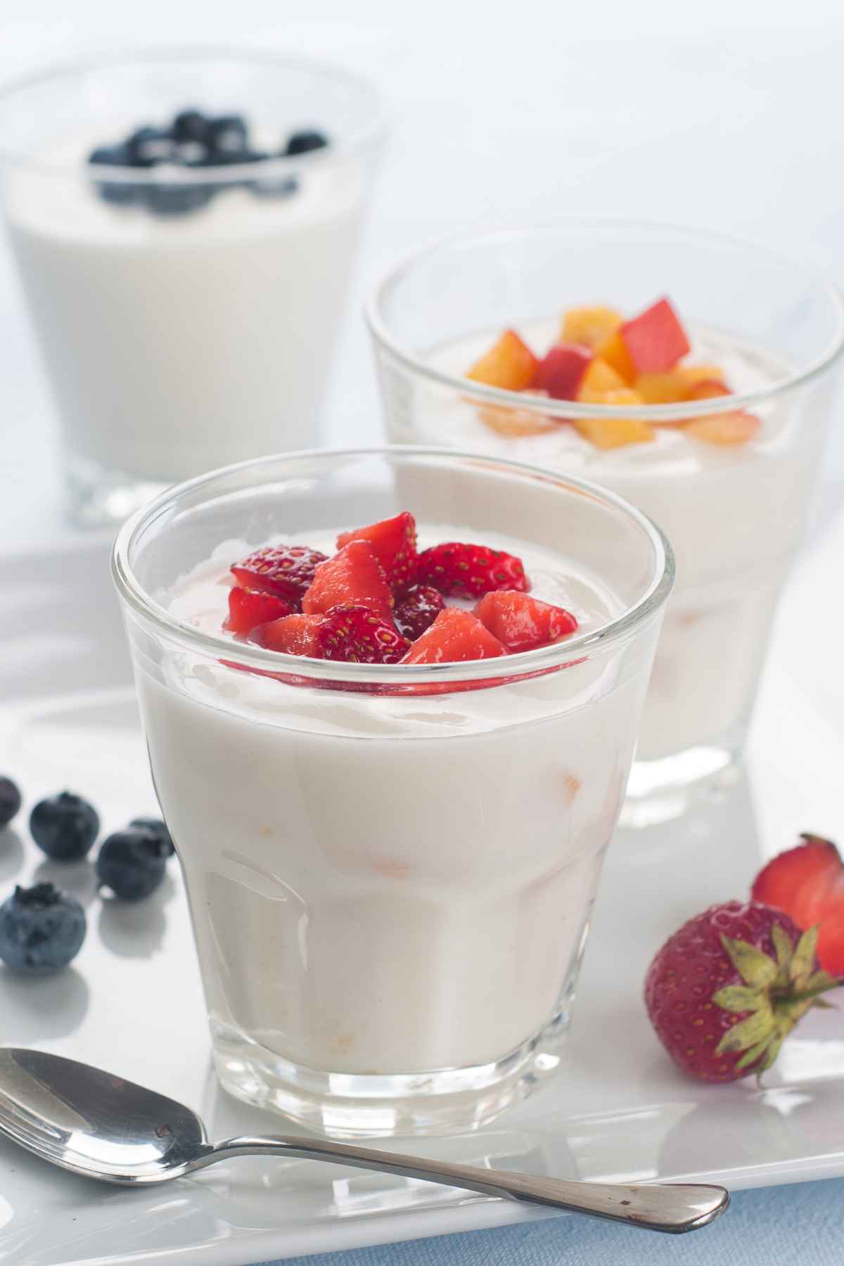 Three glasses of yogurt topped with different fruits; strawberries, peaches, and blueberries. A spoon, a few blueberries, and a strawberry are placed around the glasses.