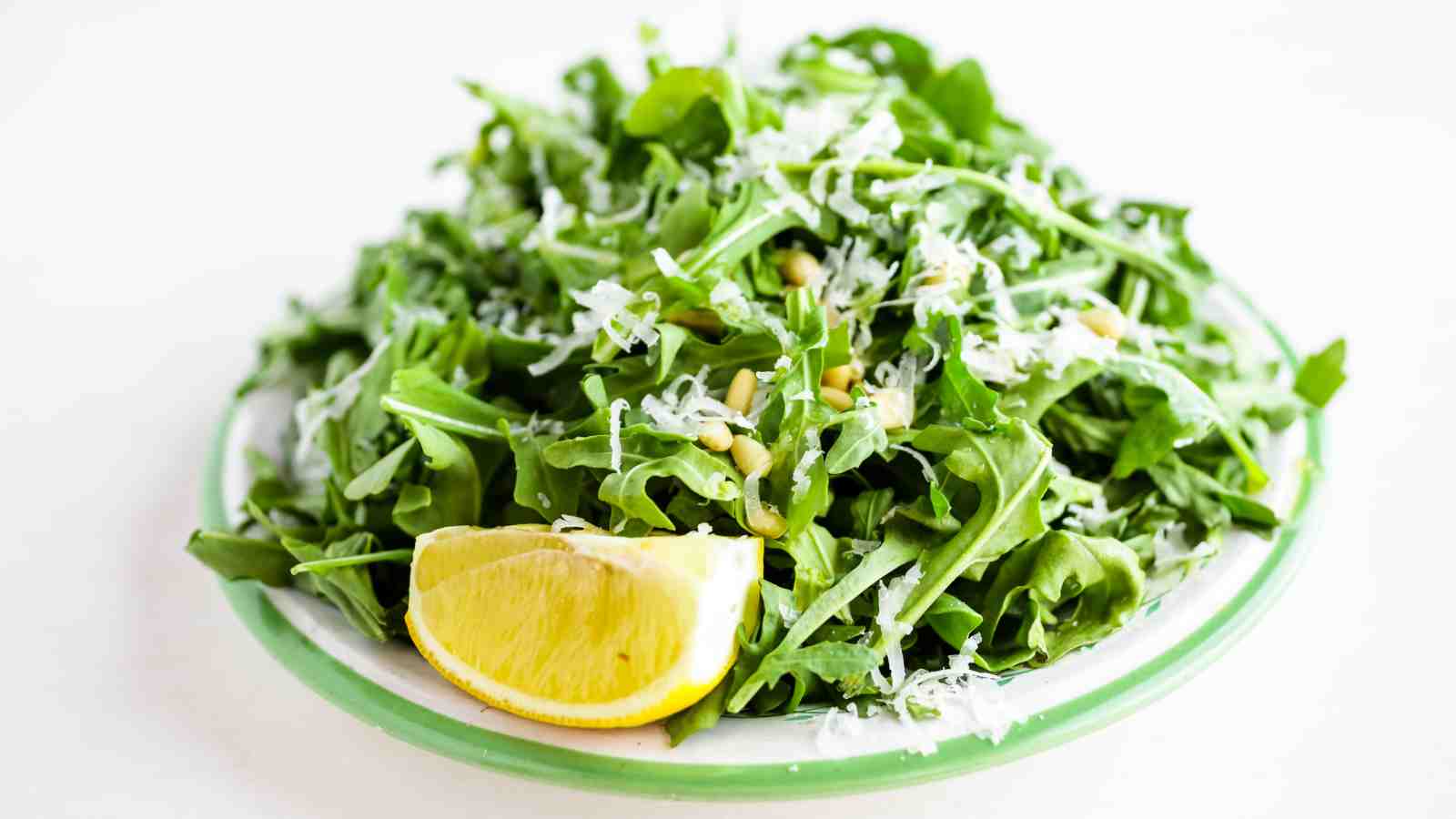 A plate of arugula salad topped with shredded cheese, pine nuts, and a lemon wedge on the side.