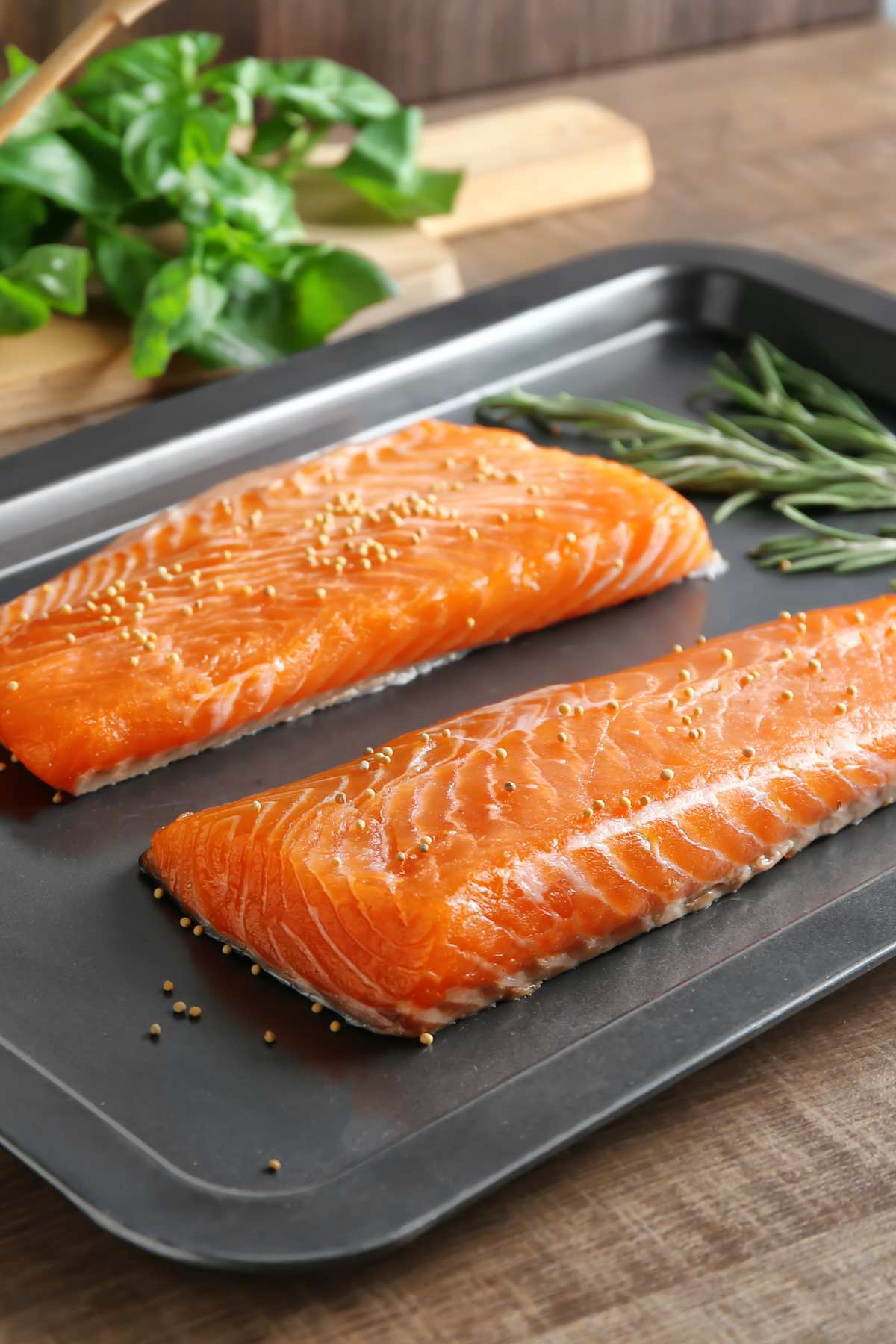 Two slices of salmon on a baking sheet beside fresh herbs on a wooden surface.