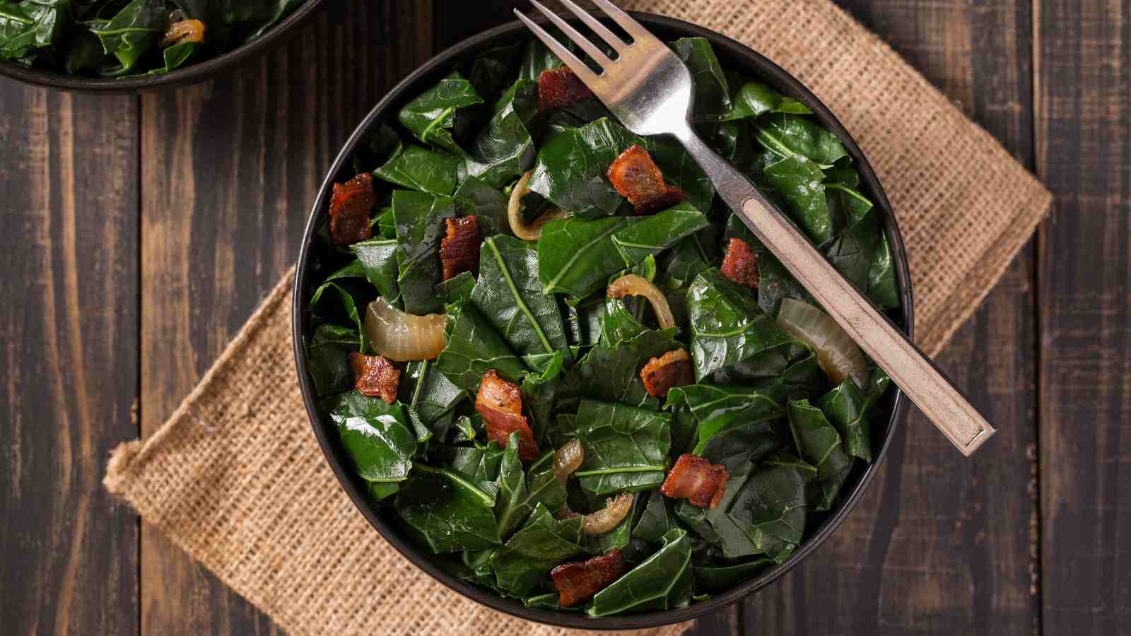 A bowl of collard greens mixed with pieces of bacon and onions, placed on a burlap mat with a fork resting on the edge.