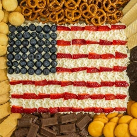 A 4th of July dessert board with an American flag design made from berries and white frosting surrounded by assorted snacks.