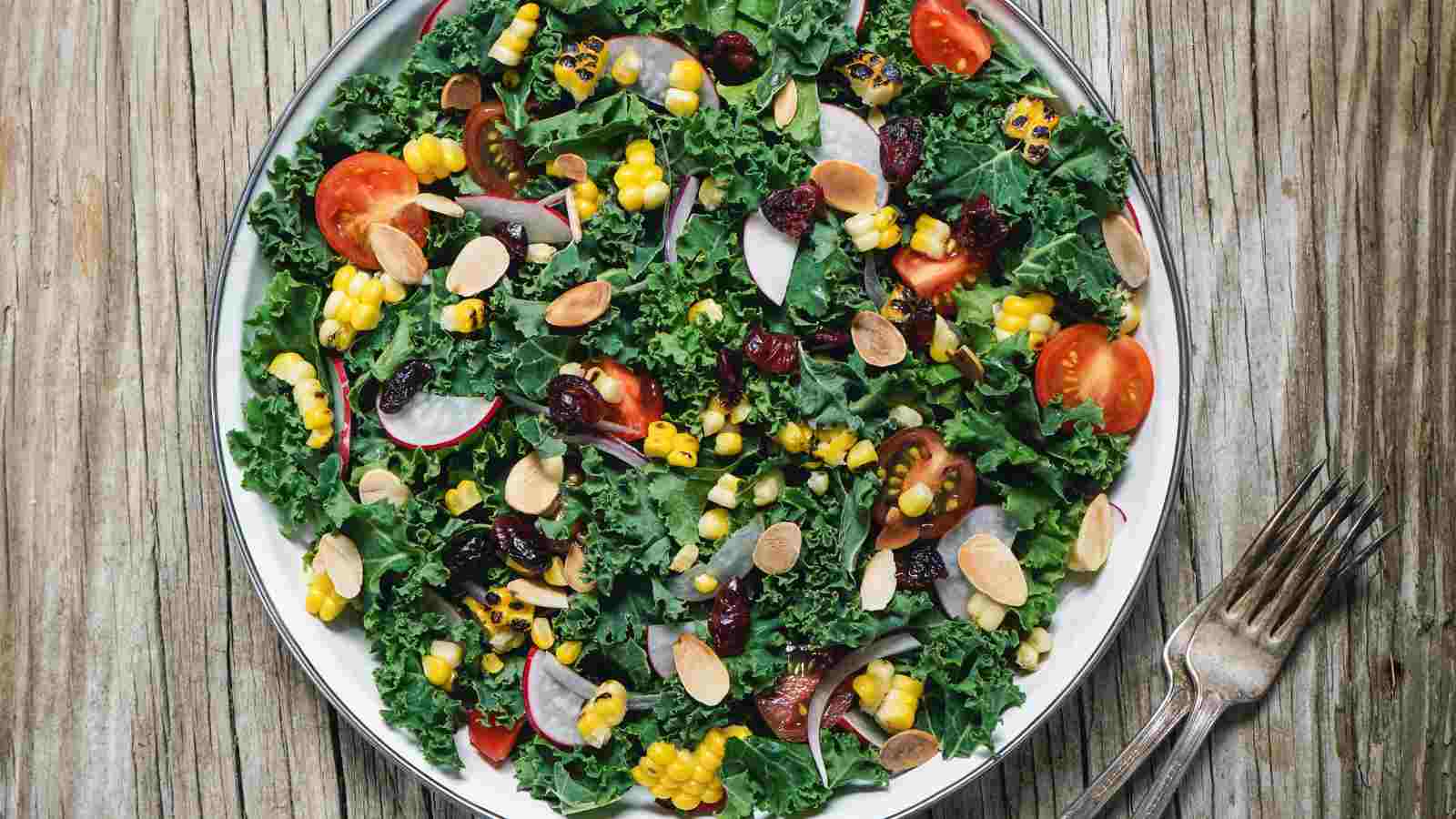 A bowl of kale salad with cherry tomatoes, sweet corn, radish slices, dried cranberries, and almond slivers.