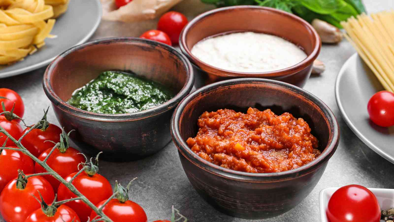 Three small bowls of pasta sauces on a wooden surface with tomatoes scattered around.