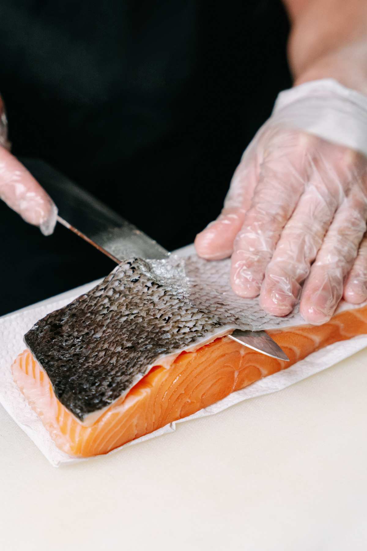 A person wearing plastic gloves is using a knife to remove the skin from a piece of raw salmon placed on a cutting board.