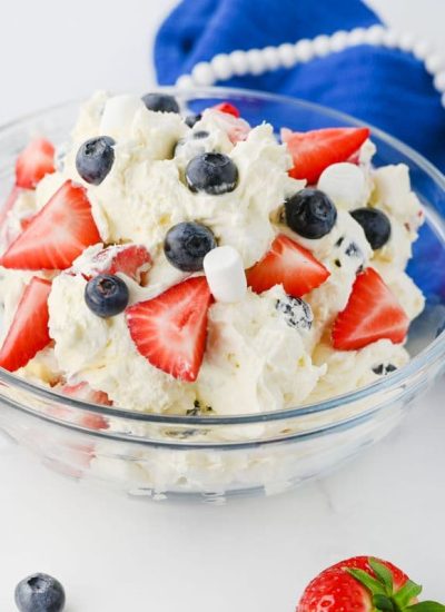 A glass bowl of patriotic dessert on a white surface, with a blue cloth and white beads in the background.