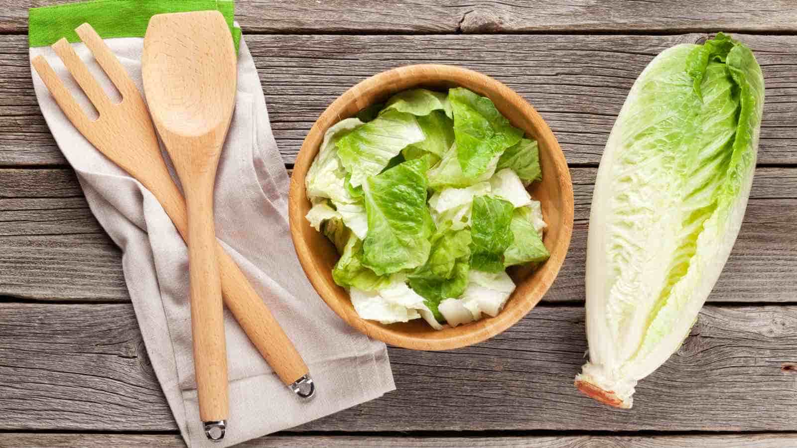 A wooden bowl with chopped lettuce, a whole romaine lettuce, and wooden salad utensils on a cloth napkin are placed on a rustic wooden surface.