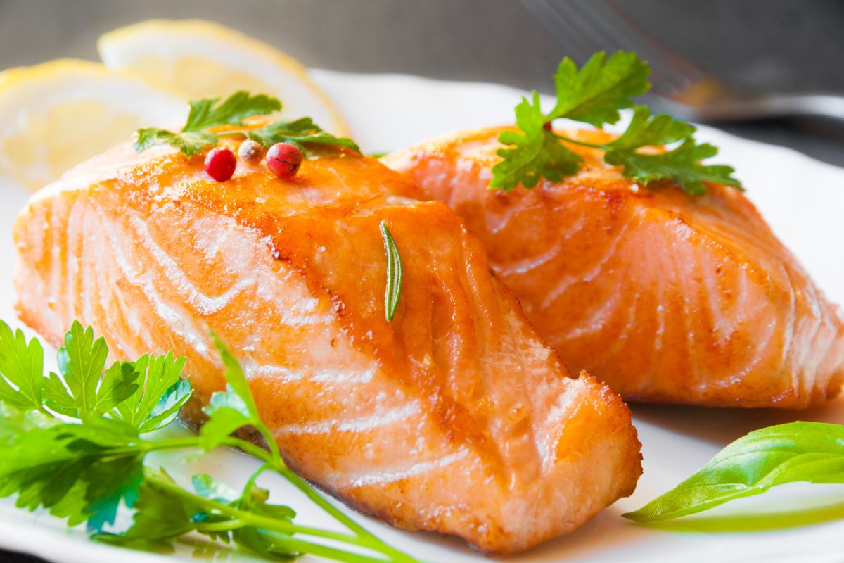 Two grilled salmon fillets garnished with sprigs of fresh parsley and pink peppercorns, served on a white plate with lemon slices in the background.