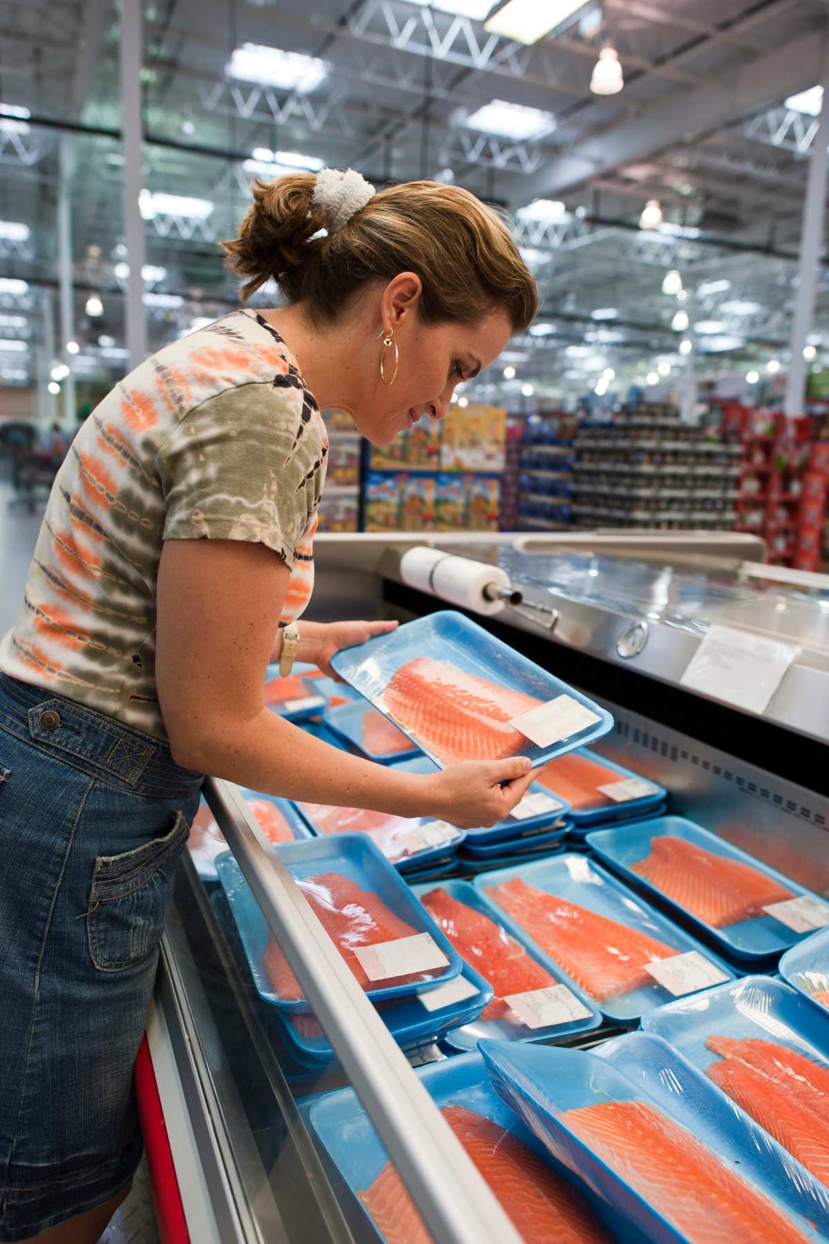 A woman is selecting packaged salmon from a refrigerated display in a grocery store.