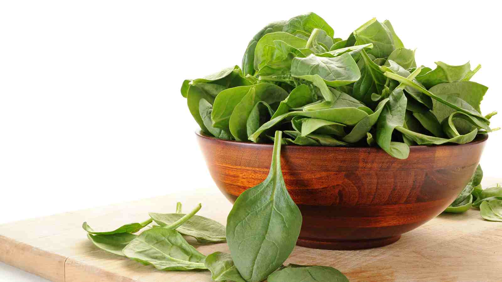 A wooden bowl filled with fresh spinach leaves sits on a light-colored wooden surface, with a few spinach leaves scattered around the bowl.
