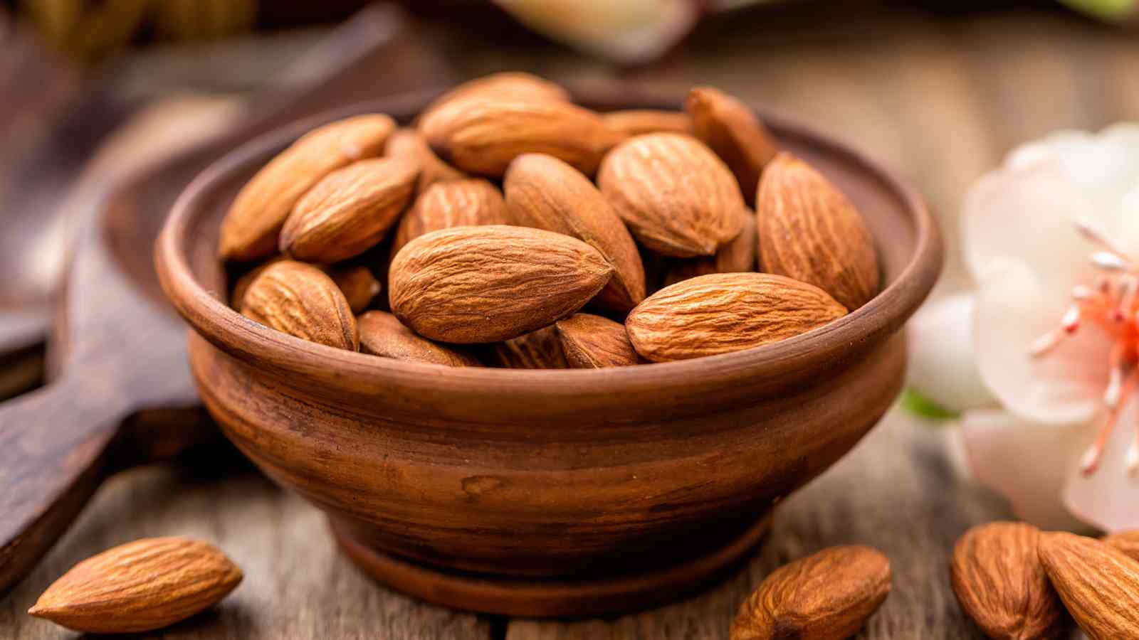 A wooden bowl filled with whole almonds, some almonds scattered around it.