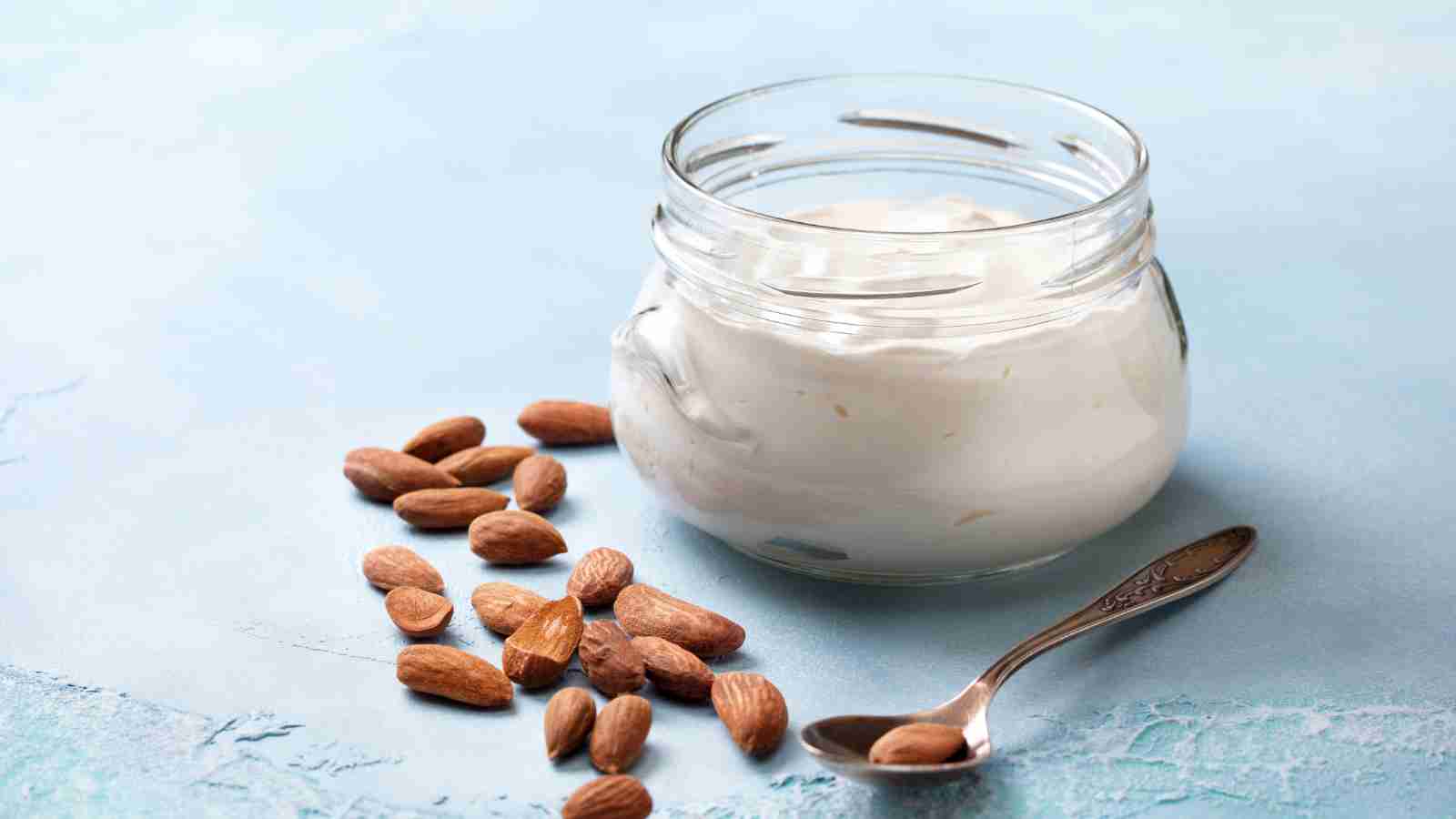 A glass jar filled with white yogurt sits on a light blue surface, with a metal spoon and a small pile of whole almonds next to it.