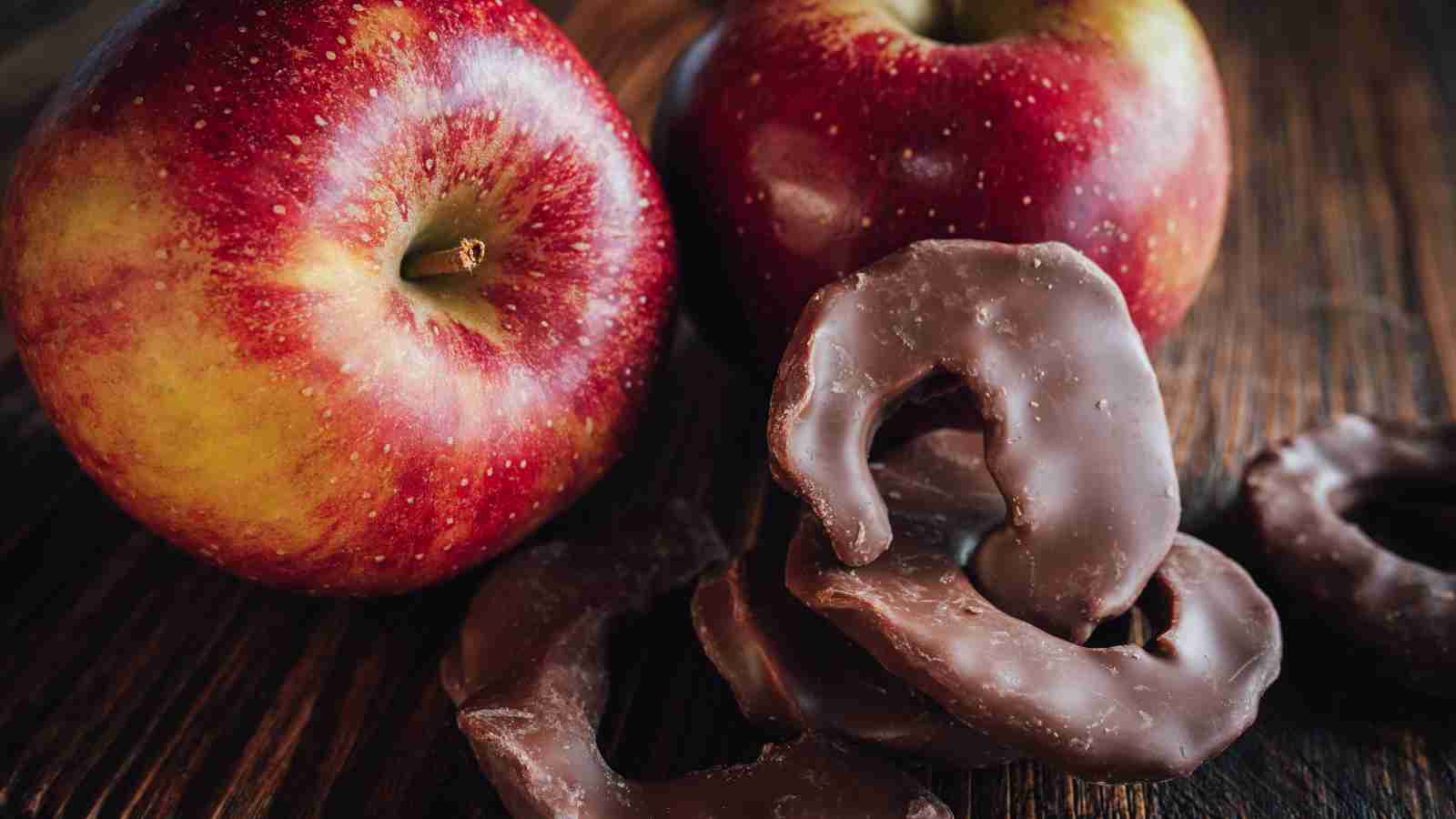 Two red apples rest on a wooden surface next to a few chocolate-covered apple slices.