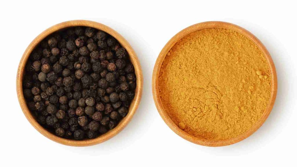 Two wooden bowls: one filled with whole black peppercorns on the left and the other with ground turmeric powder on the right, both on a white background.