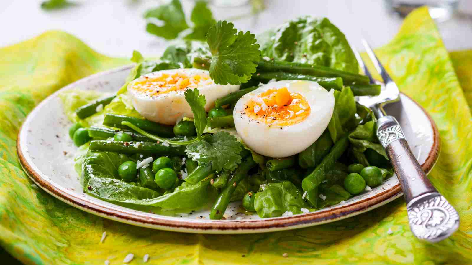 A plate of leafy green salad with two soft-boiled eggs, green beans, peas, and cilantro, garnished with black pepper.