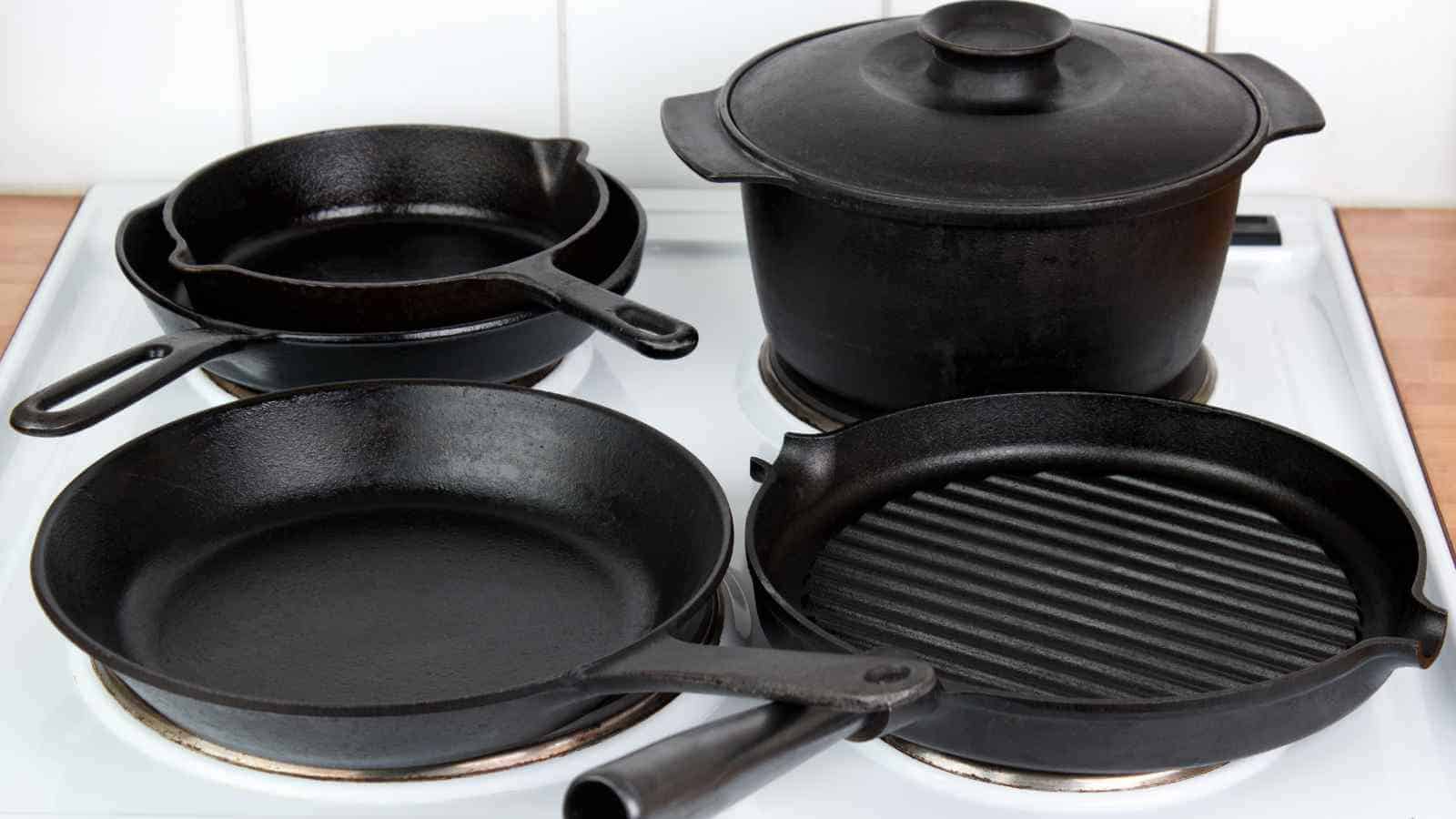 A white stove with four black cast iron cookware items: a skillet, a grill pan, a pot with a lid, and a small pan.