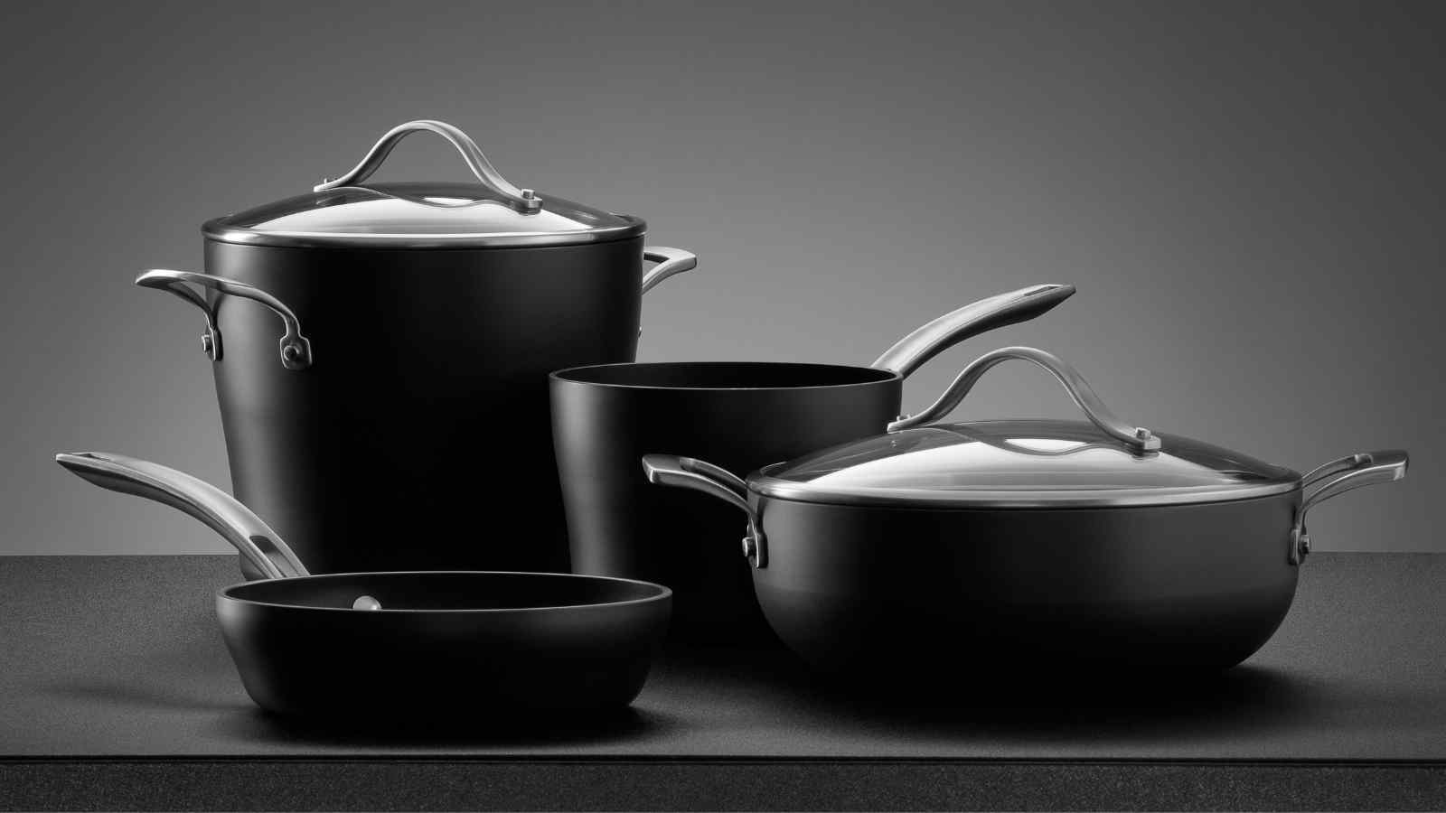A set of black ceramic cookware including a frying pan, two lidded pots, and a large lidded stew pot placed on a flat surface.