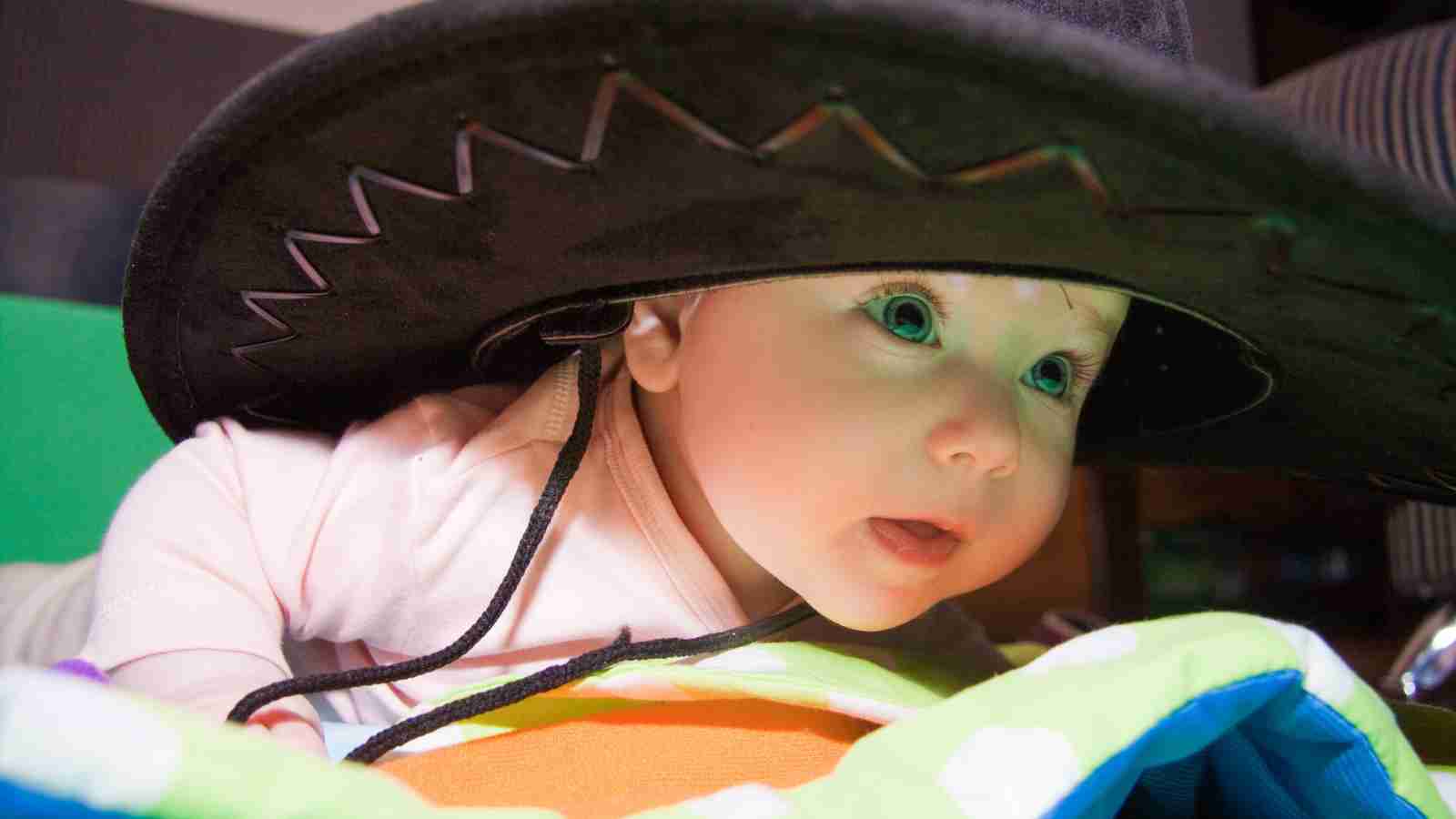 A baby wearing an oversized black hat lies on a colorful blanket, looking towards the camera with a curious expression.