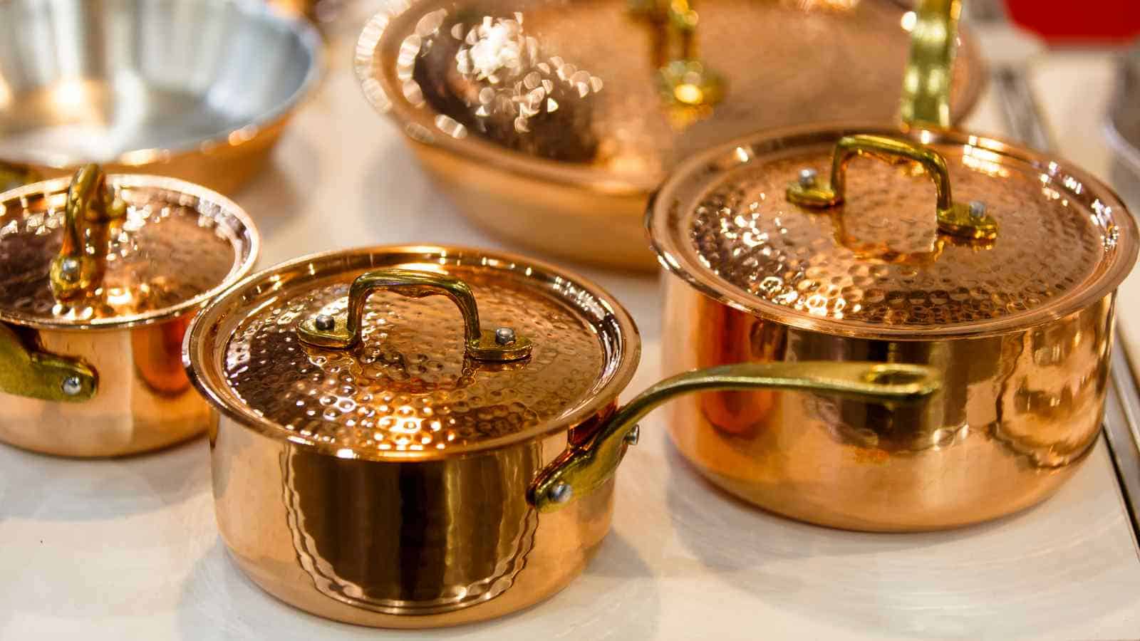 A collection of shiny copper pots and pans with lids and brass handles are displayed on a flat surface.