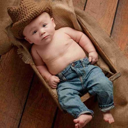 A baby wearing blue jeans and a brown knit hat lies on burlap fabric on a wooden floor.