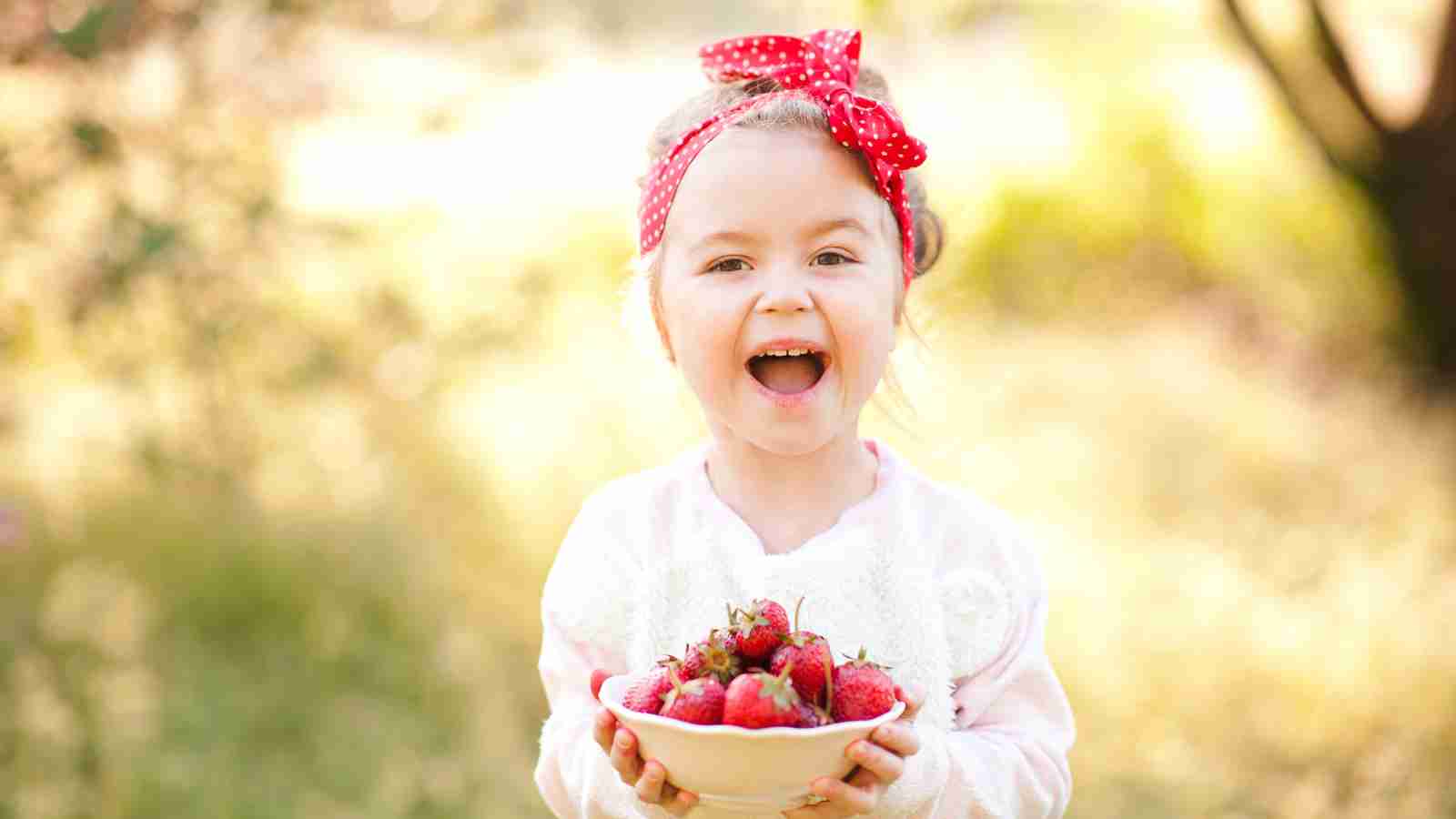 A young girl outdoors, holding a bowl filled with strawberries. She has a red headband with white polka dots and is smiling with an open mouth.