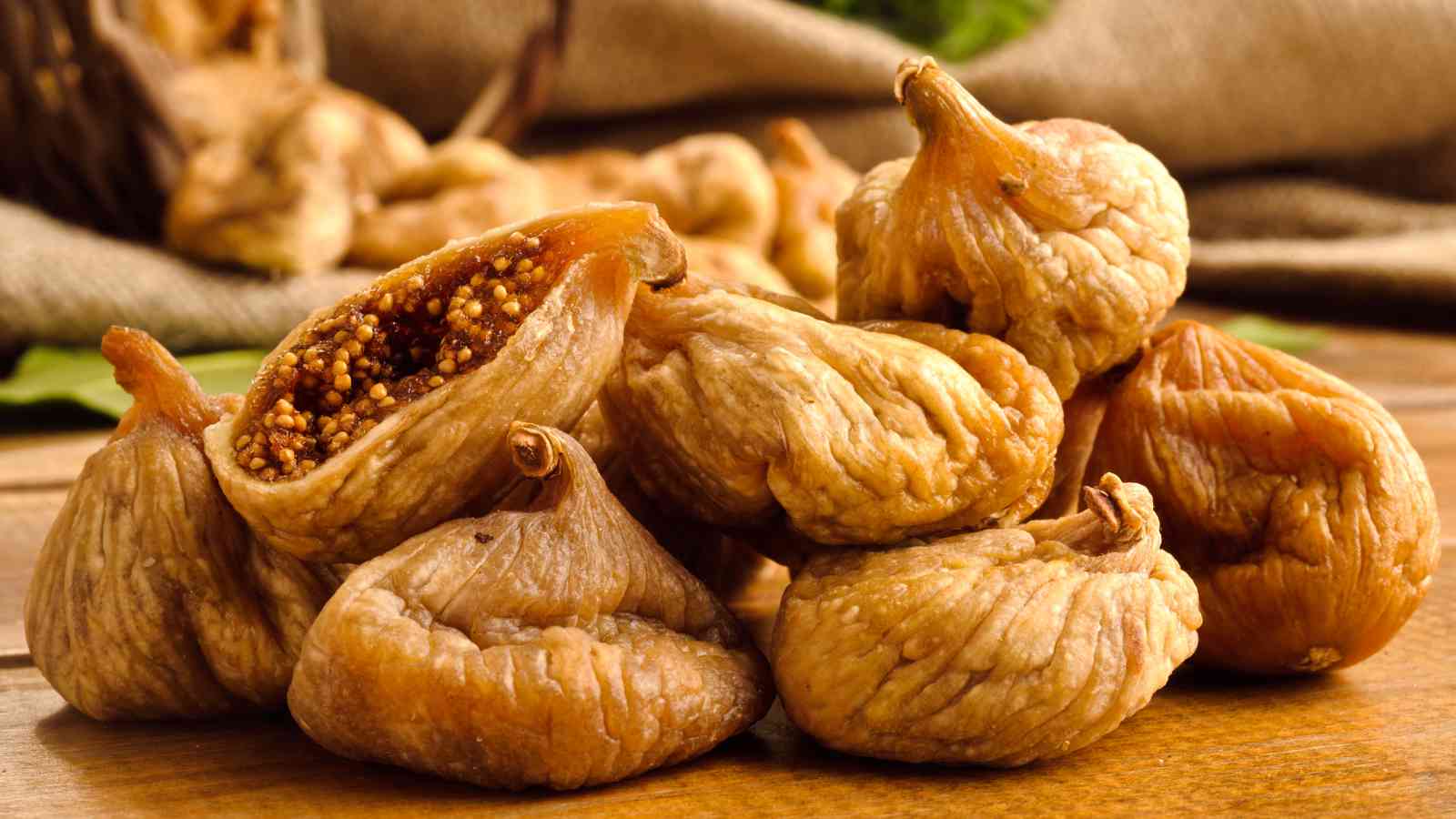 A pile of dried figs displayed on a wooden surface. Some figs are whole while others are cut open, revealing their seeds inside.