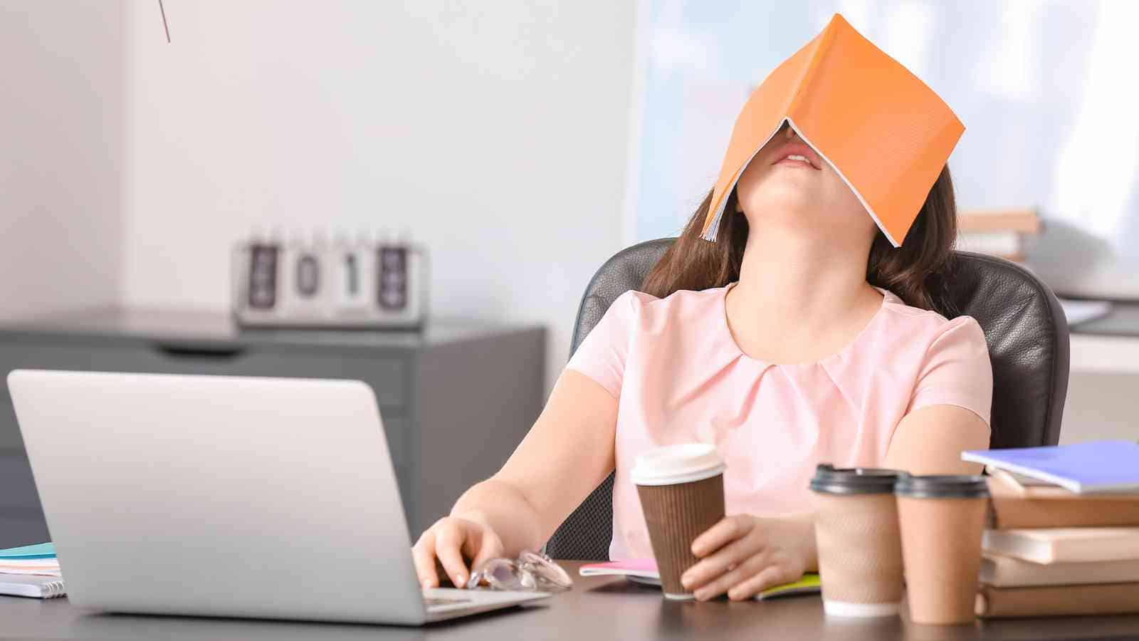 A woman sits at a desk with a laptop, holding a coffee cup, and has an open book covering her face, appearing to be asleep.