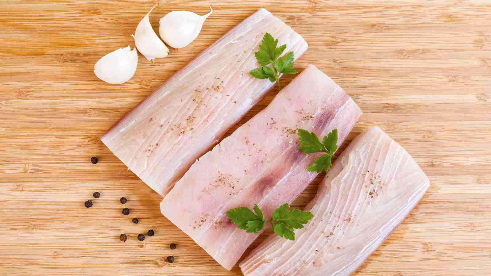 Three raw fish fillets seasoned with pepper and garnished with parsley, placed on a wooden cutting board alongside garlic cloves and black peppercorns.