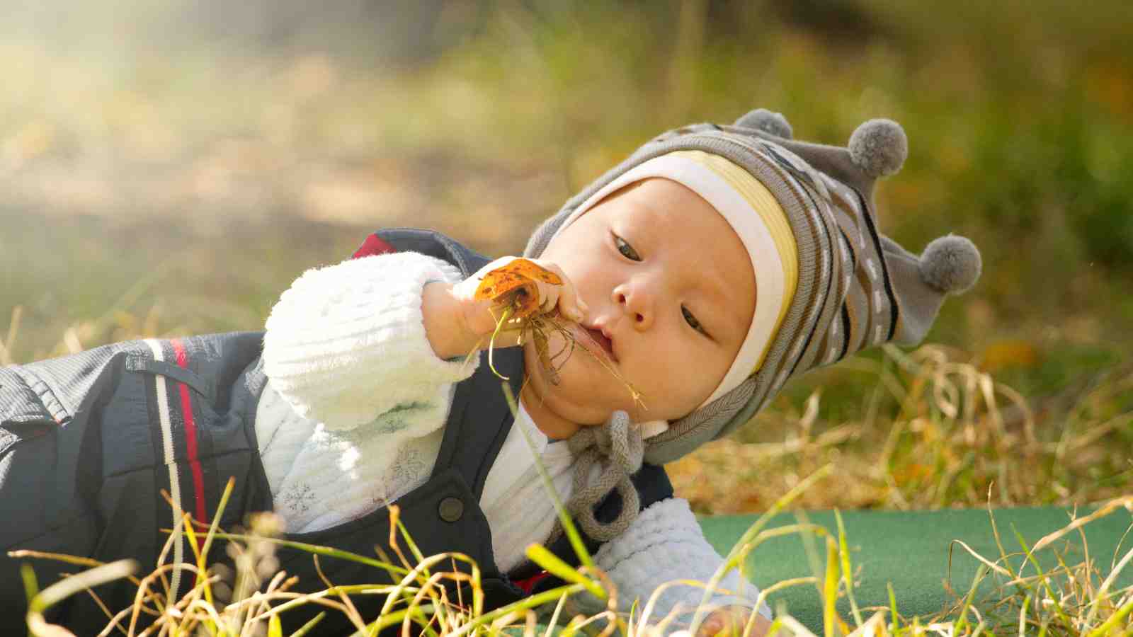 Baby lying on grass, wearing a gray beanie with pom-poms, insulated overalls, and a white sweater.