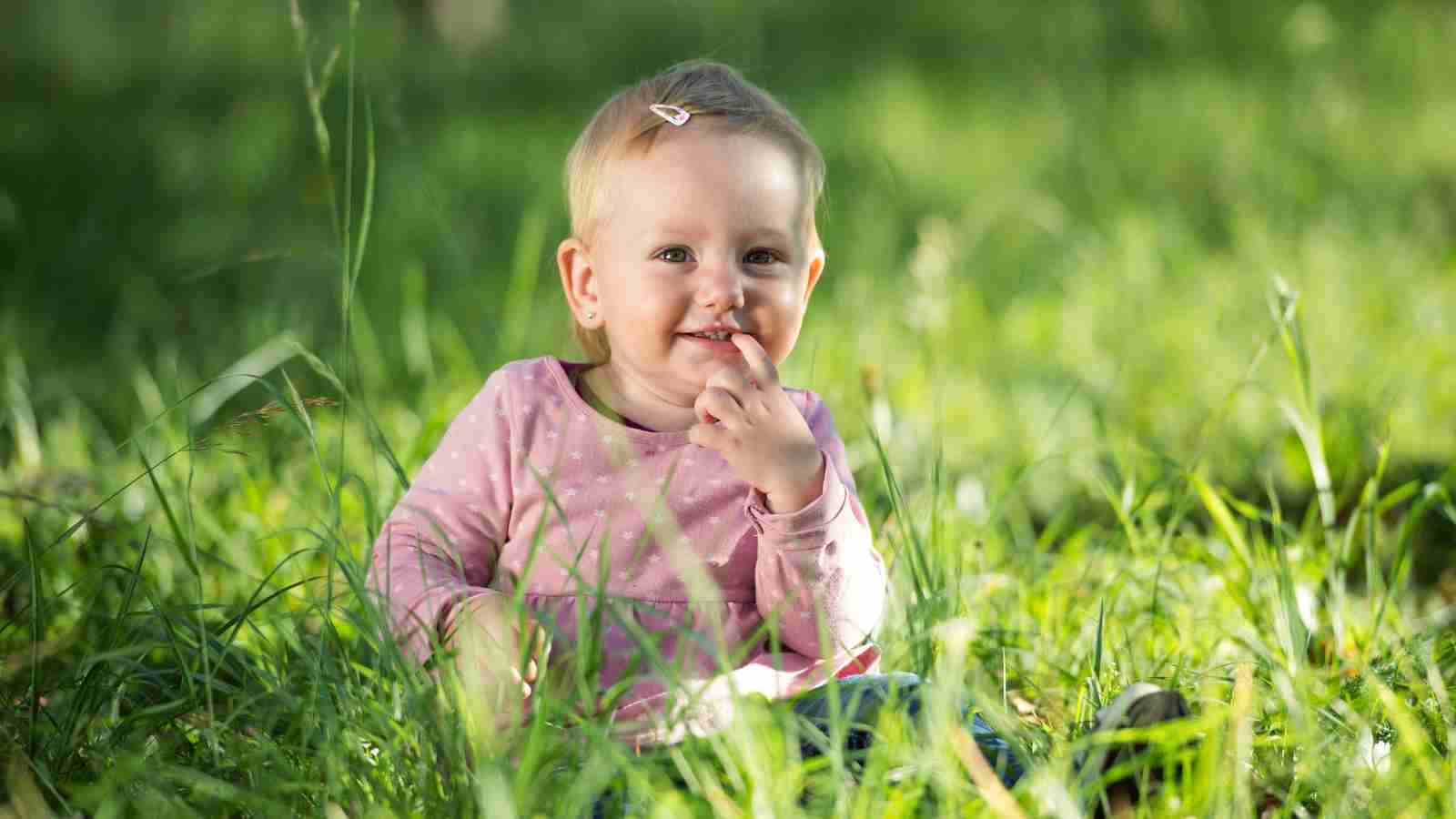 A young child with short light hair, wearing a pink shirt with white polka dots, sits in tall green grass.