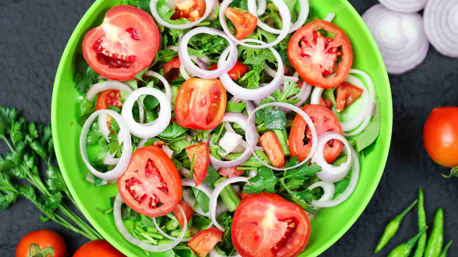 A fresh salad in a green bowl with sliced tomatoes, onion rings, cilantro, and herbs.