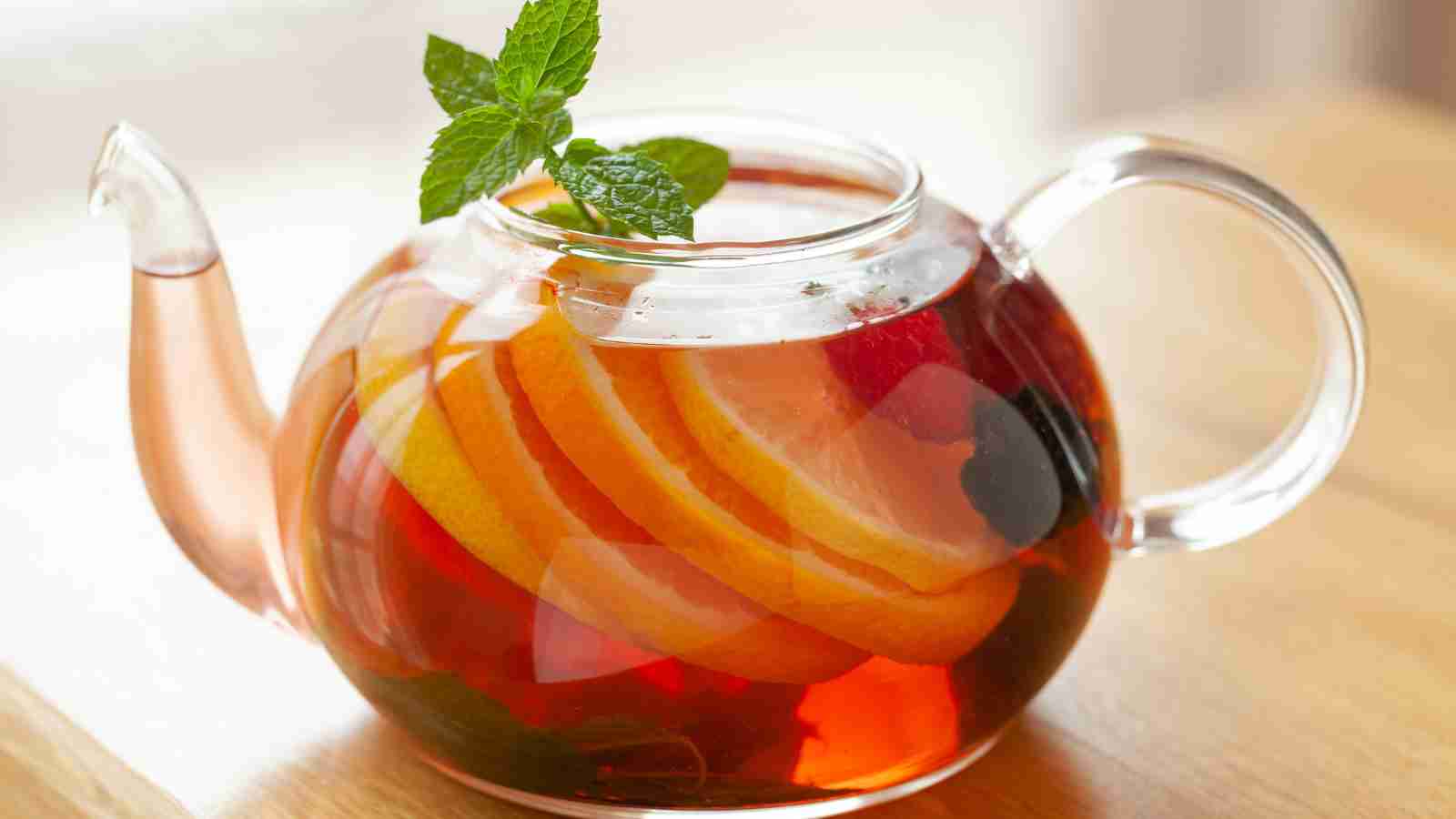 Clear glass teapot containing a red beverage with slices of citrus fruits and a sprig of mint, placed on a wooden surface.