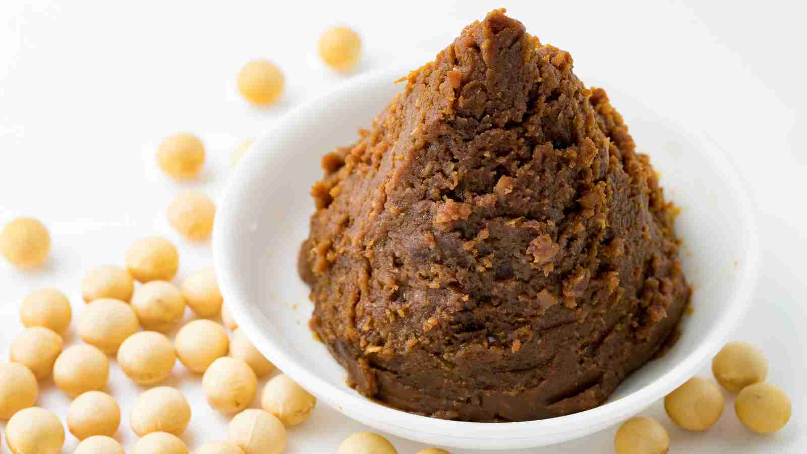 A dish of brown miso paste forming a peak in a white bowl, surrounded by scattered soybeans on a white surface.