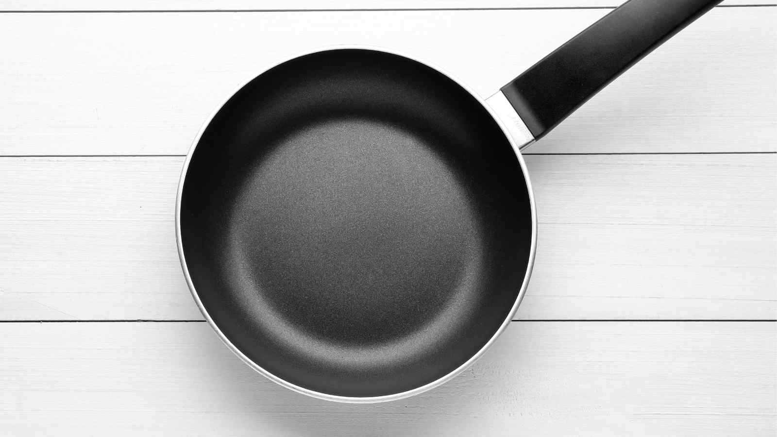 A black, non-stick frying pan with a long handle is placed on a white wooden surface.