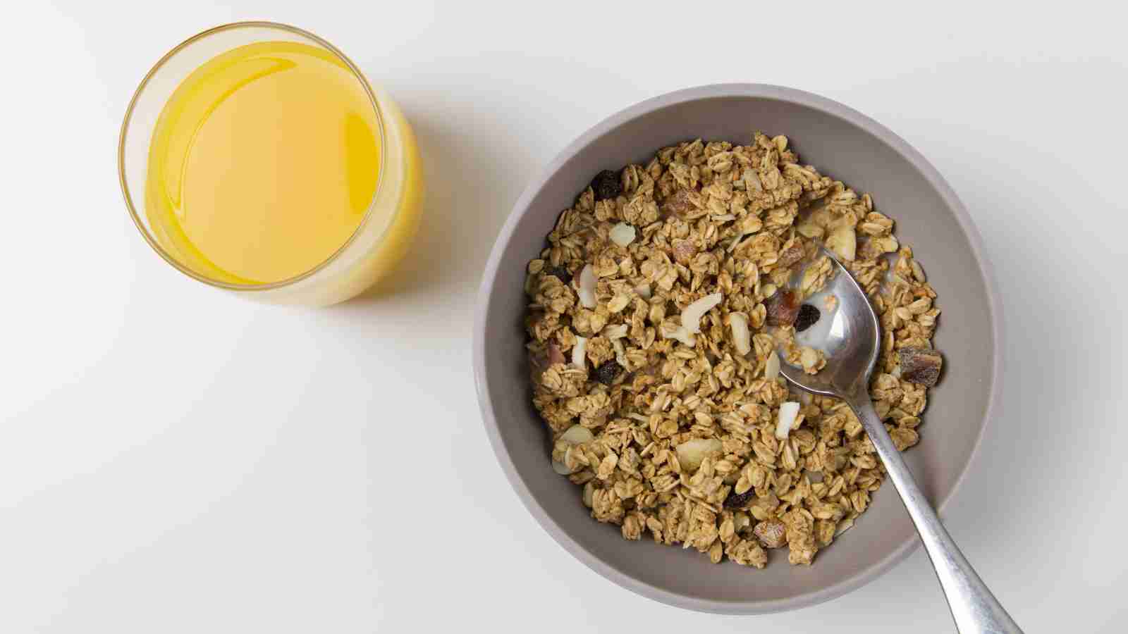 A bowl of granola with a spoon beside a glass of orange juice on a white surface.